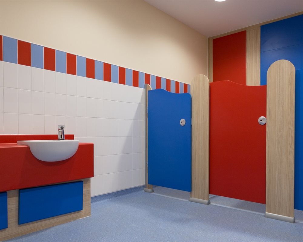 Tiny Stuff nursery toilet cubicles with blue and red doors and oak colour pilasters. Matching semi-recessed vanity unit.