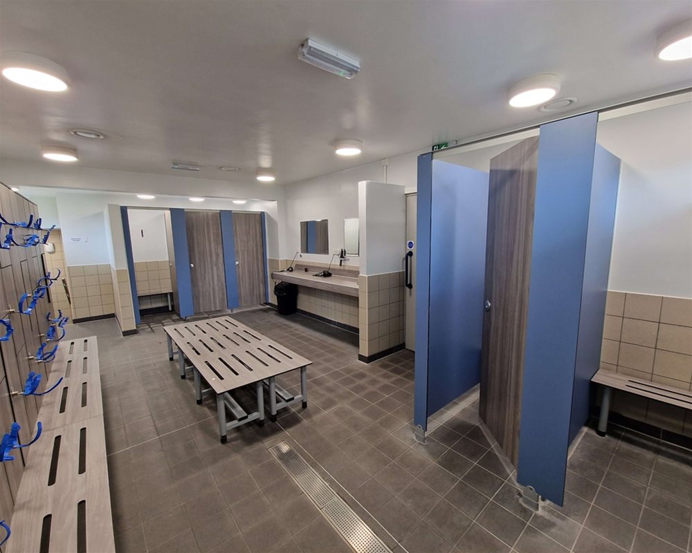 Swimming pool changing room with 'Baseline' changing cubicles in 'Grey Oak', freestanding changing room bench in the centre of the room and lockers and benching with wristband keys.