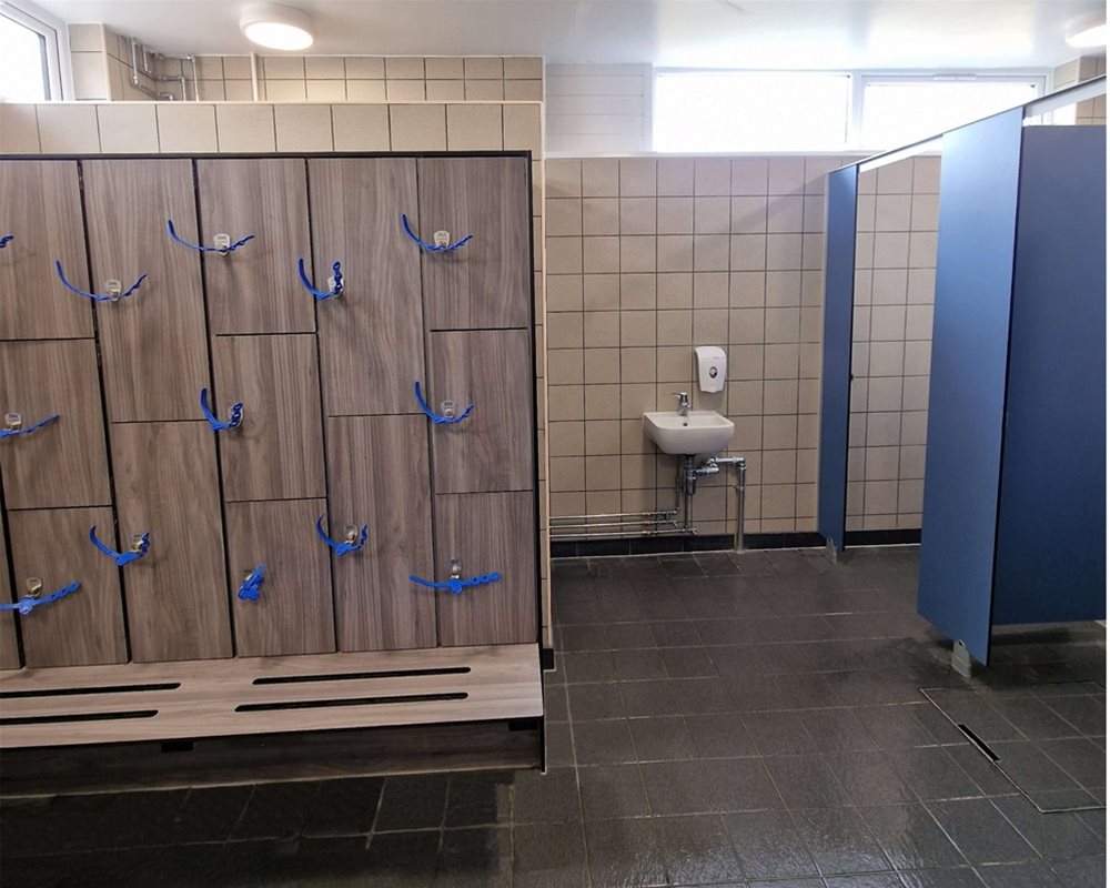 Swimming pool changing room with 'Baseline' changing cubicles in with blue pilasters and lockers and bench in 'Grey Oak' with wristband keys.