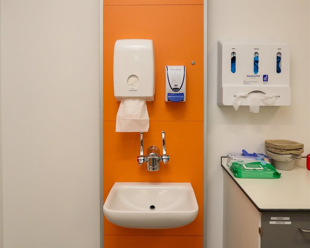 Healthcare IPS Boxed out unit in 'Orange' with clinical wash basin and wall mounted lever healthcare taps, tissue dispenser, soap dispenser in hospital room.